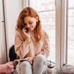 romantic red haired girl reading book beside window shy european young woman talking on phone
